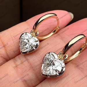 May include: Gold hoop earrings, each featuring a heart-shaped diamond pendant. The diamonds are set in silver-coloured metal and sparkle. The earrings are shown against a warm, neutral background.