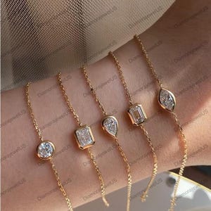 May include: A collection of gold bracelets, each featuring a different diamond shape: round, emerald cut, pear, and marquise. The diamonds are set in delicate gold bezels and connected by fine gold chains. The bracelets are displayed on a wrist.
