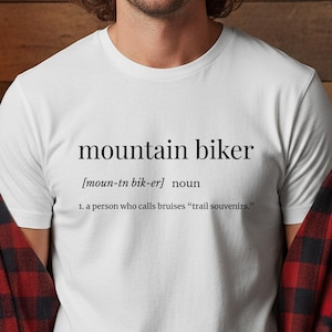May include: White t-shirt with the text "mountain biker" and its definition. The definition reads: "1. a person who calls bruises 'trail souvenirs.'" The shirt is worn with a red and black plaid shirt.