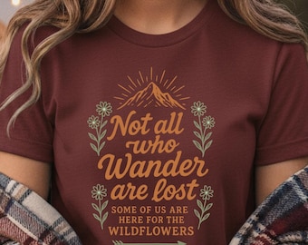 以下が含まれることがあります： 「Not all who Wander are lost」の文字が装飾的なフォントで書かれた、バーガンディ色のTシャツ。山と花の模様がデザインされています。青いジーンズを履いた人が屋外に座っています。
