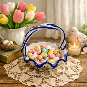 May include: A decorative ceramic basket filled with colorful Easter eggs, featuring a blue and white handle and rim. A bouquet of tulips in a white pitcher and a lit candle are in the background, creating a festive spring scene.