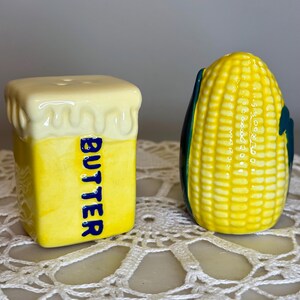 May include: A pair of ceramic salt and pepper shakers. One is shaped like a stick of butter with the word "BUTTER" in blue. The other is shaped like an ear of corn with green husks. Both are yellow and white.