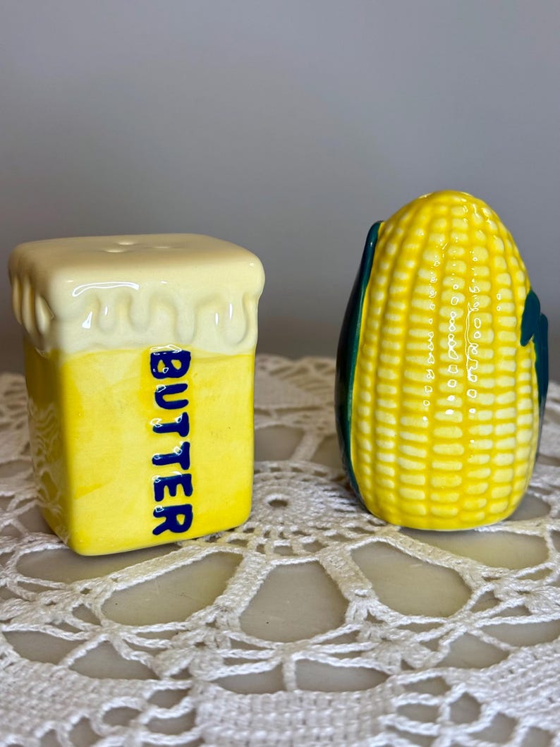 May include: A pair of ceramic salt and pepper shakers. One is shaped like a stick of butter with the word "BUTTER" in blue. The other is shaped like an ear of corn with green husks. Both are yellow and white.