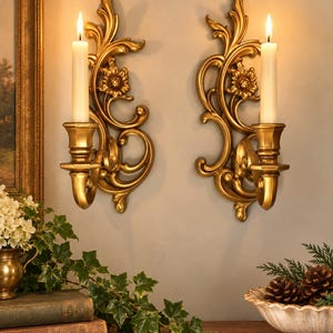 May include: A pair of gold-colored wall sconces featuring ornate scrollwork and floral motifs, each holding a single white taper candle. The sconces are mounted vertically, displaying a metallic finish with detailed carvings and curved accents.