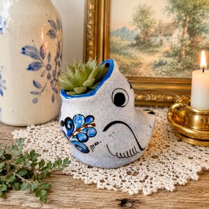 May include: A speckled light blue ceramic planter shaped like a bird, featuring hand-painted blue and brown floral accents and the signature MFX. The planter holds a small green succulent plant and has a smooth, rounded body with a wide opening.