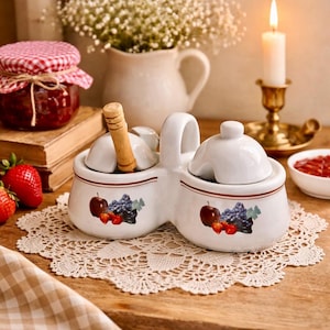 May include: A white ceramic condiment server with a handle, featuring a fruit design of apples, grapes, and strawberries. The server has two compartments, each with a small spoon and a lid. A jar of jam and fresh strawberries are also visible.