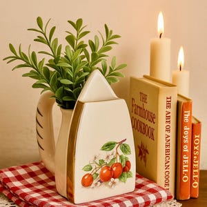 May include: A cream-colored ceramic pitcher-shaped container with a gold trim and a cherry design, holding green leafy sprigs. It sits on a red and white checkered cloth with a white lace doily, next to vintage cookbooks and lit candles.