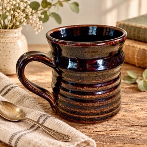 May include: A dark brown ceramic mug with a glossy finish and horizontal ribbed texture. The mug features a wide, curved handle and a flared rim. The deep brown glaze shows subtle variations in tone, highlighting the ridges along the body.