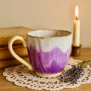 May include: A ceramic mug with a purple floral design, a cream-colored handle, and a light green rim. The mug sits on a white lace doily, with a lit candle and a bundle of lavender in the background. The mug is approximately 4 inches tall.