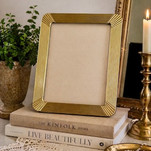 May include: A gold-colored rectangular picture frame with a ribbed design, resting on a stack of books. The books have the titles "THE KINFOLK HOME" and "LIVE BEAUTIFULLY". A lit candle in a gold candlestick and a small decorative tray are also visible.