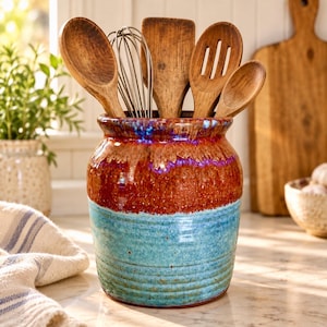 May include: A cylindrical ceramic utensil crock featuring a two-tone glaze with a deep reddish-brown top and a textured light blue base. The crock is filled with various wooden kitchen spoons, a spatula, and a metal wire whisk.
