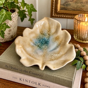 May include: A ceramic dish shaped like an oak leaf with a glossy cream-colored glaze, featuring a prominent crackle finish and a central splash of blue and green hues. The edges are wavy and organic, mimicking the natural form of a fallen leaf.