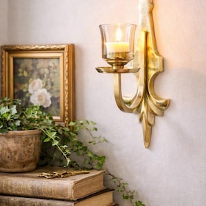 May include: A wall-mounted candle holder with a lit candle in a glass cup. The holder is made of gold-colored metal and has an ornate design. A framed floral painting, a potted plant, and a stack of books are also visible.