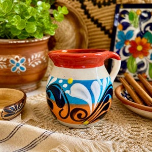 May include: A hand-painted ceramic pitcher with a white base, adorned with blue, orange, and black floral designs. The rim is red, and it has a white handle. The pitcher is on a textured surface, with other decorative items in the background.