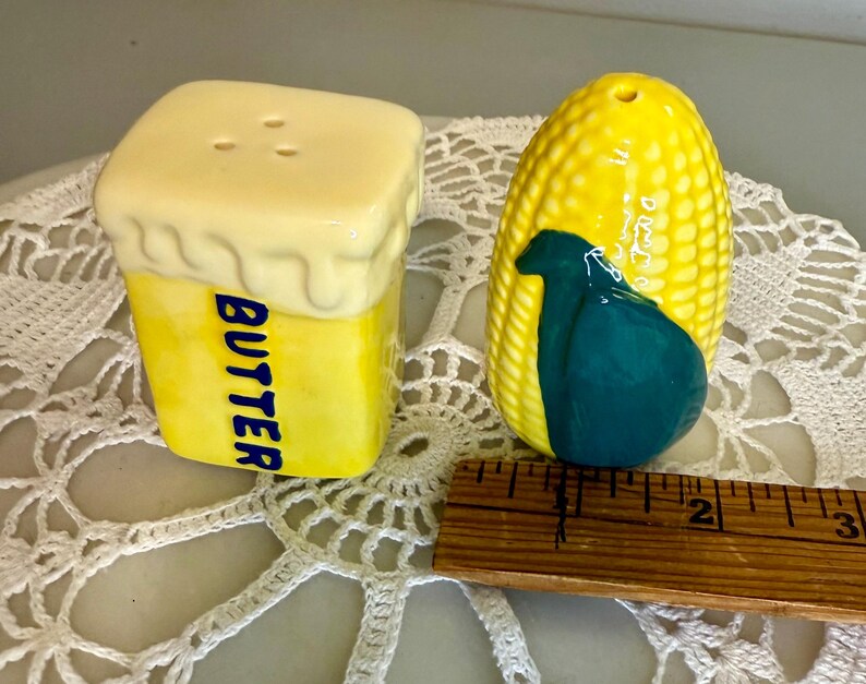 May include: A pair of ceramic salt and pepper shakers. One is shaped like a yellow butter stick with the word "BUTTER" in blue. The other is a yellow corn cob with a teal design.