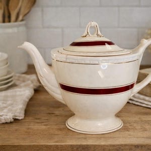 May include: Cream-colored teapot with a lid, featuring a wide, dark red stripe and gold trim. The teapot has a curved spout and handle, and sits on a pedestal base. The background includes other kitchenware and a wooden surface.