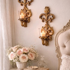 May include: Two ornate gold wall sconces with glass candle holders, each holding a lit candle. A bouquet of pink roses and baby's breath sits on a bedside table with books, pearls, and a perfume bottle. The scene evokes a romantic, vintage aesthetic.