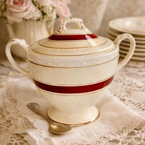 May include: Cream-colored sugar bowl with a lid, featuring a wide red band and gold trim. The bowl has two handles and sits on a pedestal base. A small silver spoon rests on a lace doily. The text "Homer Laughlin Georgian Eggshell" is at the bottom.
