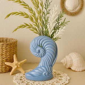 May include: A light blue ceramic seashell-shaped vase holds green and white faux plants. A starfish, a woven basket, a seashell, and a seashell wreath decorate the scene. The vase sits on a cream-colored doily.
