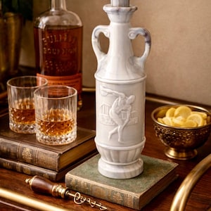 May include: A white ceramic decanter with a relief design of a figure throwing a discus, flanked by two handles. The decanter is on a wooden bar cart with a bottle of bourbon, two glasses of whiskey, and a bowl of lemon slices.