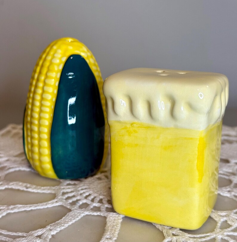 May include: A pair of ceramic salt and pepper shakers. One is shaped like a yellow ear of corn with a dark teal side. The other is a yellow square with a white foam top, resembling a beer mug. Both have small holes for dispensing.