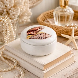 May include: A white ceramic trinket box with a lid featuring a seashell design. The box sits atop two off-white books. A pearl necklace, a glass of coral, and a perfume bottle are in the background.