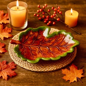 May include: A ceramic leaf-shaped serving dish with a brown interior and green trim, resting on a woven coaster. Two lit candles and fall foliage complete the autumn-themed display.
