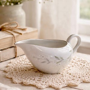 May include: White porcelain gravy boat with a delicate floral pattern and a silver rim. The gravy boat sits on a cream-colored crocheted doily. A small handle is on the side. The background includes books and a vase of flowers.