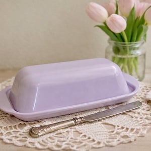 May include: A light purple butter dish with a matching lid sits on a cream-colored doily. A silver butter knife rests beside the dish. A small glass jar with pink tulips and a folded linen napkin are in the background.