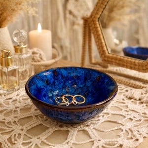 May include: A deep blue ceramic bowl with gold rings inside sits on a cream-colored lace doily. The bowl has a textured, glazed finish. Other items on the wooden surface include a woven basket, perfume bottles, a candle, and a decorative mirror.