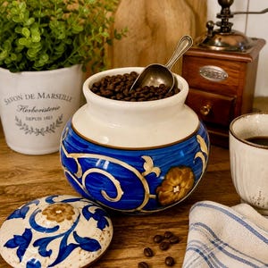 May include: A round ceramic jar with a cream-colored top and a vibrant blue base featuring hand-painted gold floral motifs. The jar is filled with dark roasted coffee beans and includes a small metal scoop. A matching lid with blue and gold patterns sits beside the jar on a wooden surface.