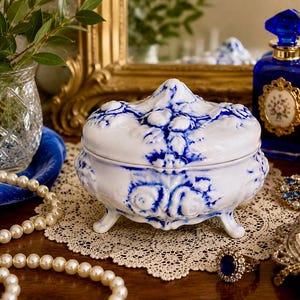 May include: A white and blue porcelain trinket box with a floral design. The box has three small feet and a decorative lid. The image also includes a blue perfume bottle, a pearl necklace, and a lit candle.