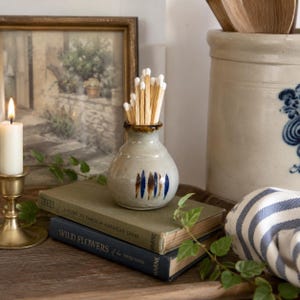 May include: A small, light gray ceramic vase with a narrow neck and a hand-painted pattern of vertical blue and brown dashes. The vase is filled with a cluster of wooden matches with white tips, resting on top of two stacked vintage hardcover books.