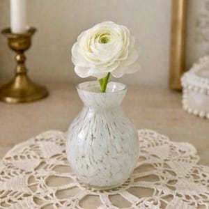 May include: A white speckled glass vase holds a single white ranunculus flower. The vase sits on a cream-colored lace doily. A gold candlestick and a decorative box are in the background, creating a soft, elegant aesthetic.