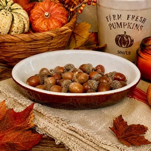 May include: A shallow, oval-shaped ceramic bowl with a dark red exterior and a white interior, filled with a collection of brown acorns. The bowl sits on a textured, light-colored fabric cloth, surrounded by autumn-themed decorative items.