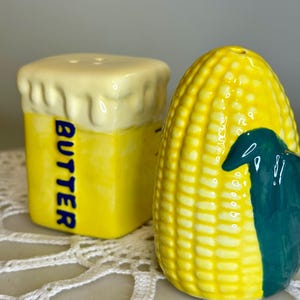 May include: A pair of ceramic salt and pepper shakers. One is shaped like a stick of butter with the word "BUTTER" in blue. The other is a yellow corn on the cob with a green husk. Both have holes for dispensing.