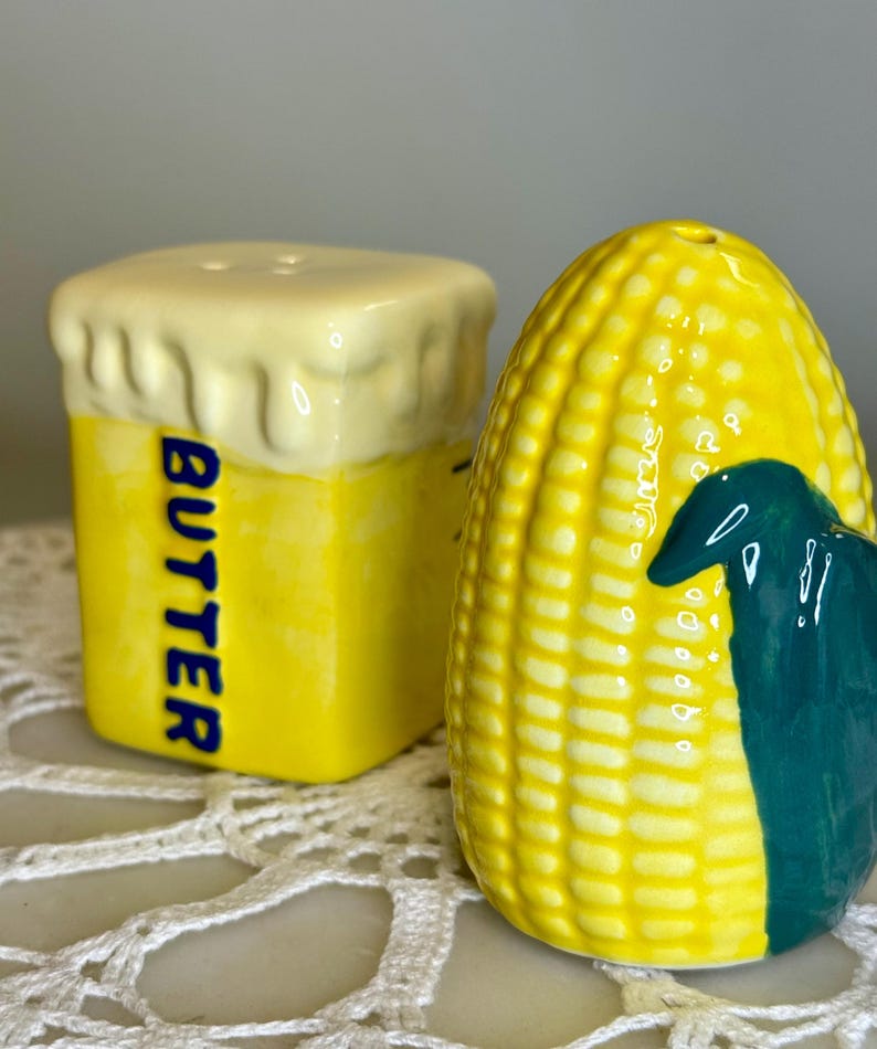 May include: A pair of ceramic salt and pepper shakers. One is shaped like a stick of butter with the word "BUTTER" in blue. The other is a yellow corn on the cob with a green husk. Both have holes for dispensing.