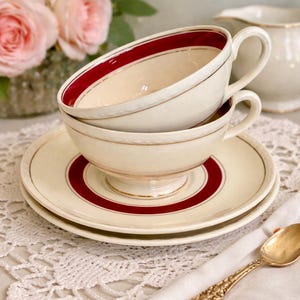 May include: Cream-colored teacups and saucers with a red band and gold trim are stacked. A gold spoon and a white napkin are on the table. Pink roses and a creamer are in the background. The teacups are vintage.