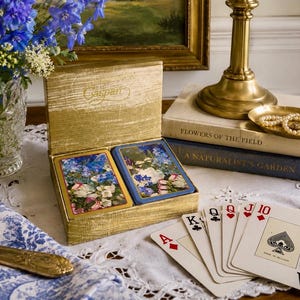 May include: A gold-colored box containing two decks of playing cards with floral designs, featuring the brand name Caspari. A hand of five playing cards is fanned out, showing the Ace of Diamonds, King of Clubs, Queen of Hearts, Jack of Hearts, and Ten of Diamonds.