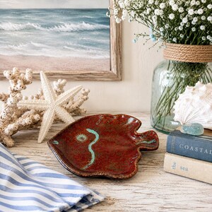 May include: A fish-shaped ceramic dish with a glossy, mottled red glaze and a single turquoise line accent. The dish features a textured surface with a small, circular turquoise detail near the head and a defined tail section on the right side.
