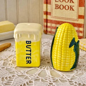May include: A yellow ceramic butter dish and a corn-shaped salt shaker sit on a white lace doily. The butter dish is rectangular with the word "BUTTER" in blue. A cookbook is in the background.
