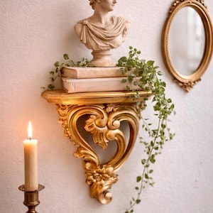 May include: A gold-colored, ornate wall shelf with a bust of a classical figure, books, and trailing greenery. A lit candle in a brass holder and a gold-framed oval mirror complete the decorative arrangement.