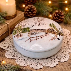 May include: A round, white ceramic trinket box with a hand-painted winter scene. The lid features a snowy landscape with a wooden fence, trees, and a small cabin. The box sits on a lace doily.