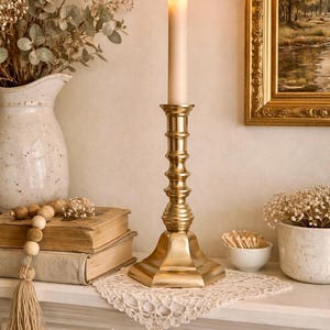 May include: A lit ivory candle sits in a gold candlestick with a square base. The candlestick is on a white surface with a lace doily. A vase of flowers, books, and a framed painting are in the background.
