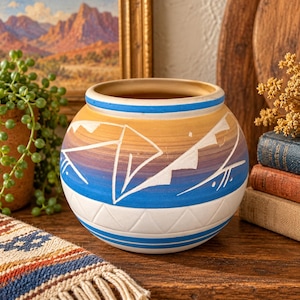 May include: A round ceramic pot featuring a gradient color scheme transitioning from yellow to purple and blue. The surface is decorated with white etched geometric mountain-like patterns and horizontal lines, with a solid white base section.