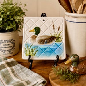 May include: A square white ceramic tile featuring a raised relief of a mallard duck swimming in blue water with green reeds and brown cattails. The tile has a diamond-patterned background and a small hole at the top for hanging on a wall.