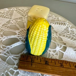 May include: A ceramic salt and pepper shaker set. The salt shaker is a yellow block with three holes. The pepper shaker is a yellow corn on the cob with green husks. The set is on a white lace doily.
