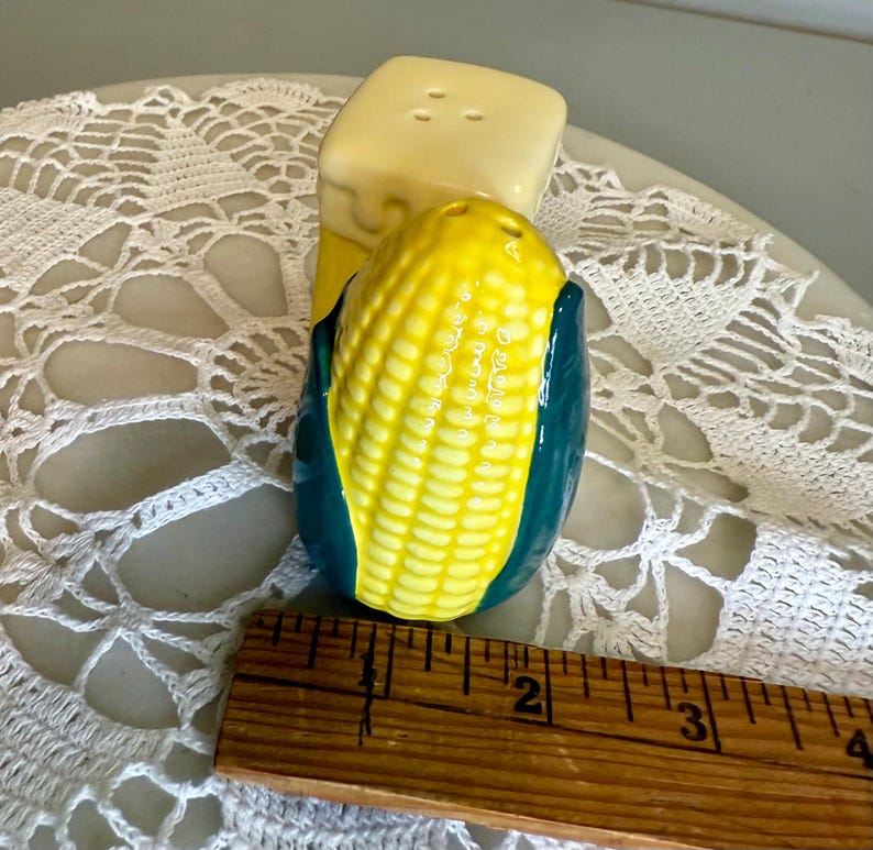 May include: A ceramic salt and pepper shaker set. The salt shaker is a yellow block with three holes. The pepper shaker is a yellow corn on the cob with green husks. The set is on a white lace doily.