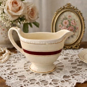 May include: Cream-colored gravy boat with a handle and spout, featuring a burgundy stripe and gold trim. The boat sits on a white lace doily, with a floral arrangement and a framed picture in the background.