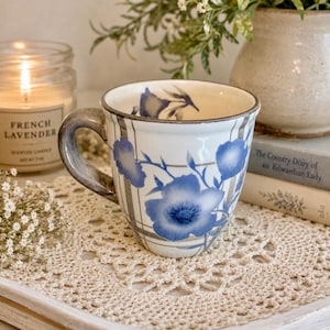 May include: A ceramic mug with a blue floral design and a gray handle. The mug is white with blue flowers and gray vertical stripes. It sits on a white tray with a crocheted doily.